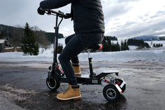 Person riding a scooter on a snowy landscape with trees and mountains in the background