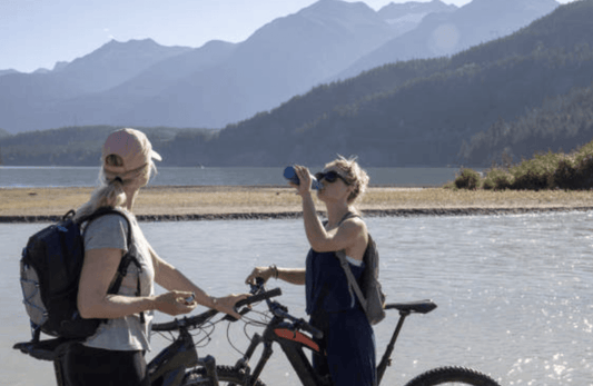 Two women with electric bikes by a lake, mountains in background, outdoor adventure scene