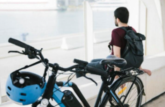 Electric bike with blue helmet in foreground, man with backpack seated by waterfront